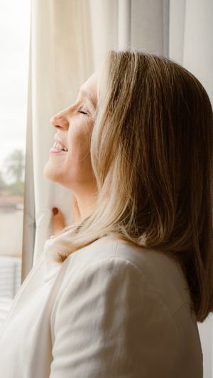 A senior woman enjoys a peaceful moment with eyes closed by a sunlit window indoors.
